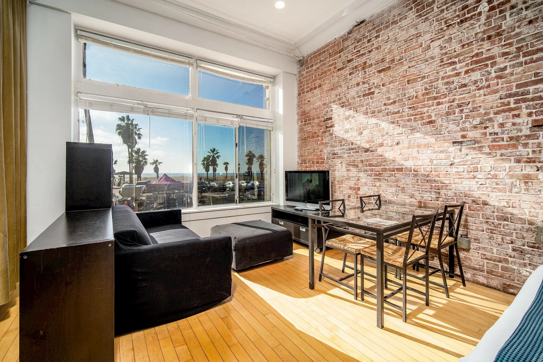 Sala de estar con pared decorativa de ladrillo, ventanas grandes, sofá, televisor y mesa de comedor.