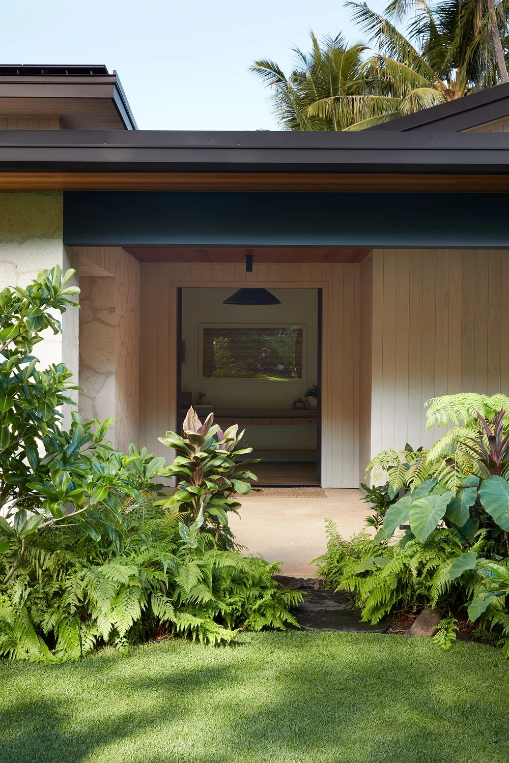 A modern house entrance opens onto a covered area, surrounded by tropical plants.