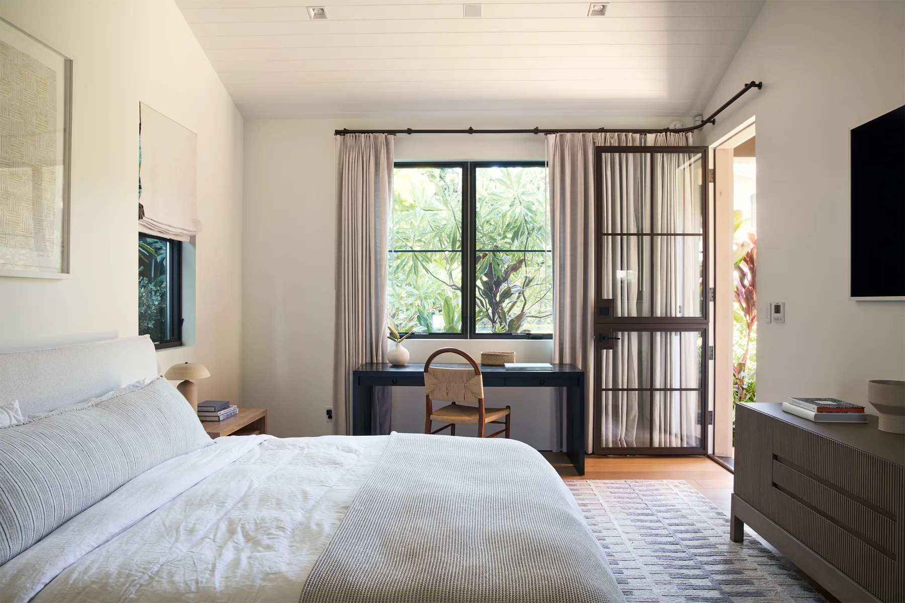 A bright bedroom with a bed, desk, and large window overlooking greenery.
