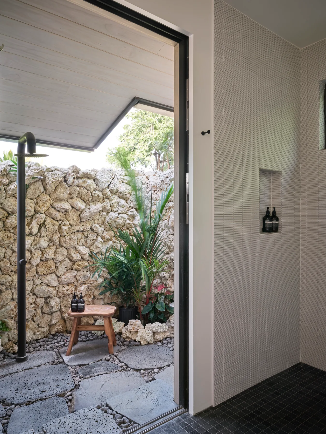 A tiled shower area opens onto a small outdoor patio with plants and a stone wall.