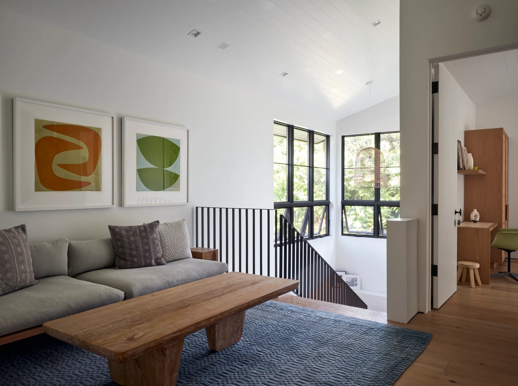 A bright living area with seating, framed artwork, and large windows overlooking a staircase.