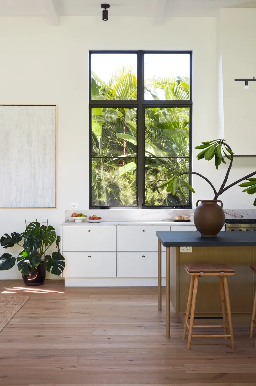 A bright kitchen with white cabinets, a large window, and plants.