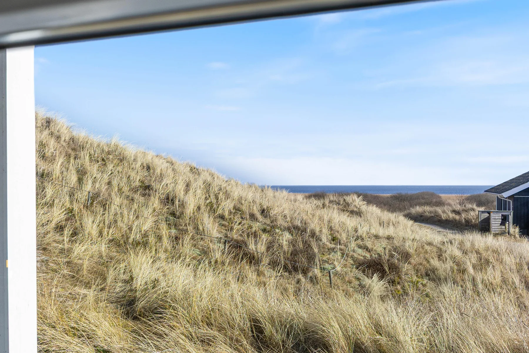 Vista de dunas cubiertas de hierba hacia el mar, enmarcada por una ventana abierta.