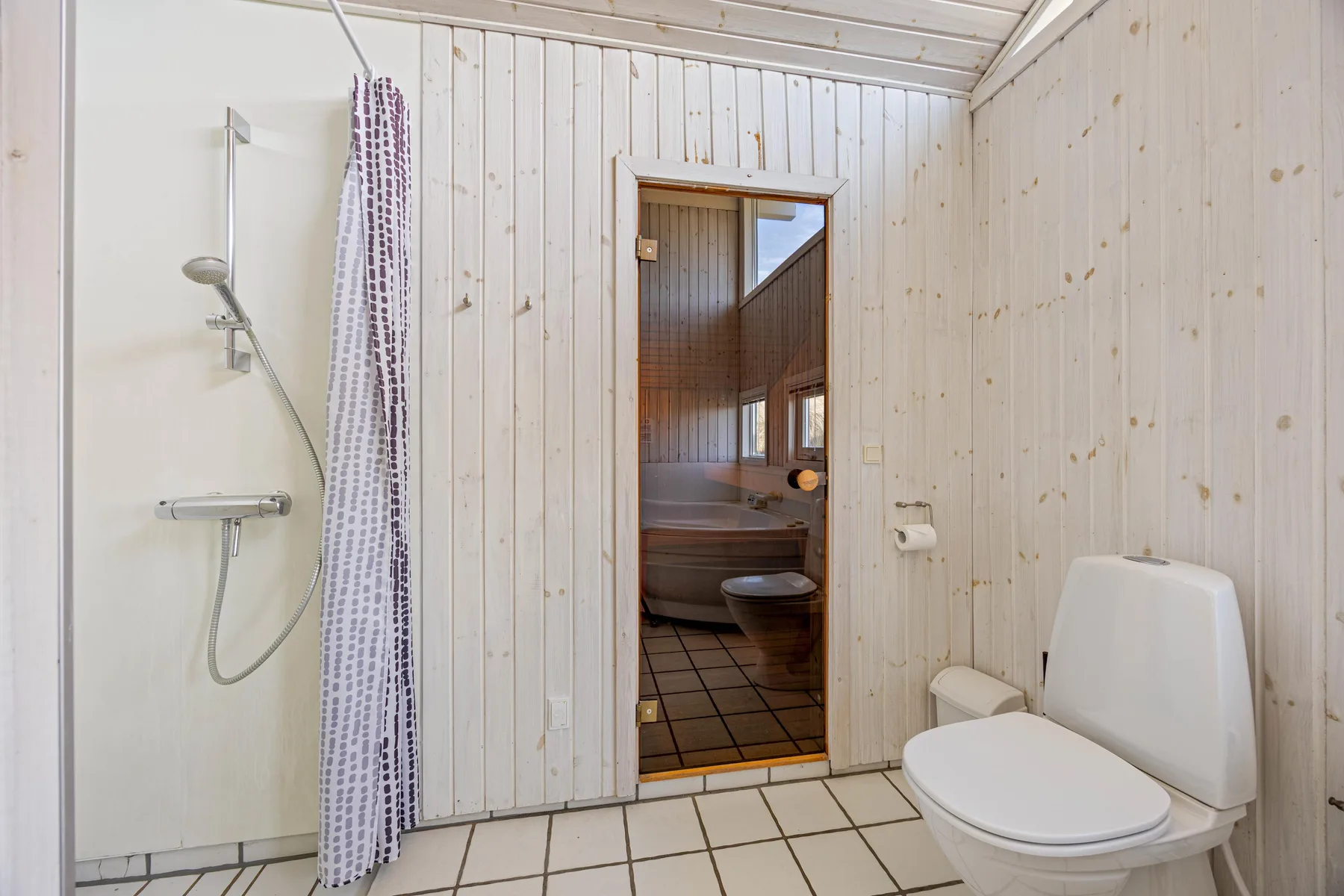 Un baño de baldosas con ducha e inodoro, con una sauna visible a través de la puerta.