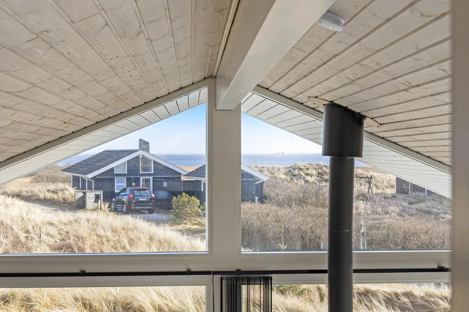 Una ventana amplia da a dunas cubiertas de hierba y una casa oscura bajo un techo inclinado.