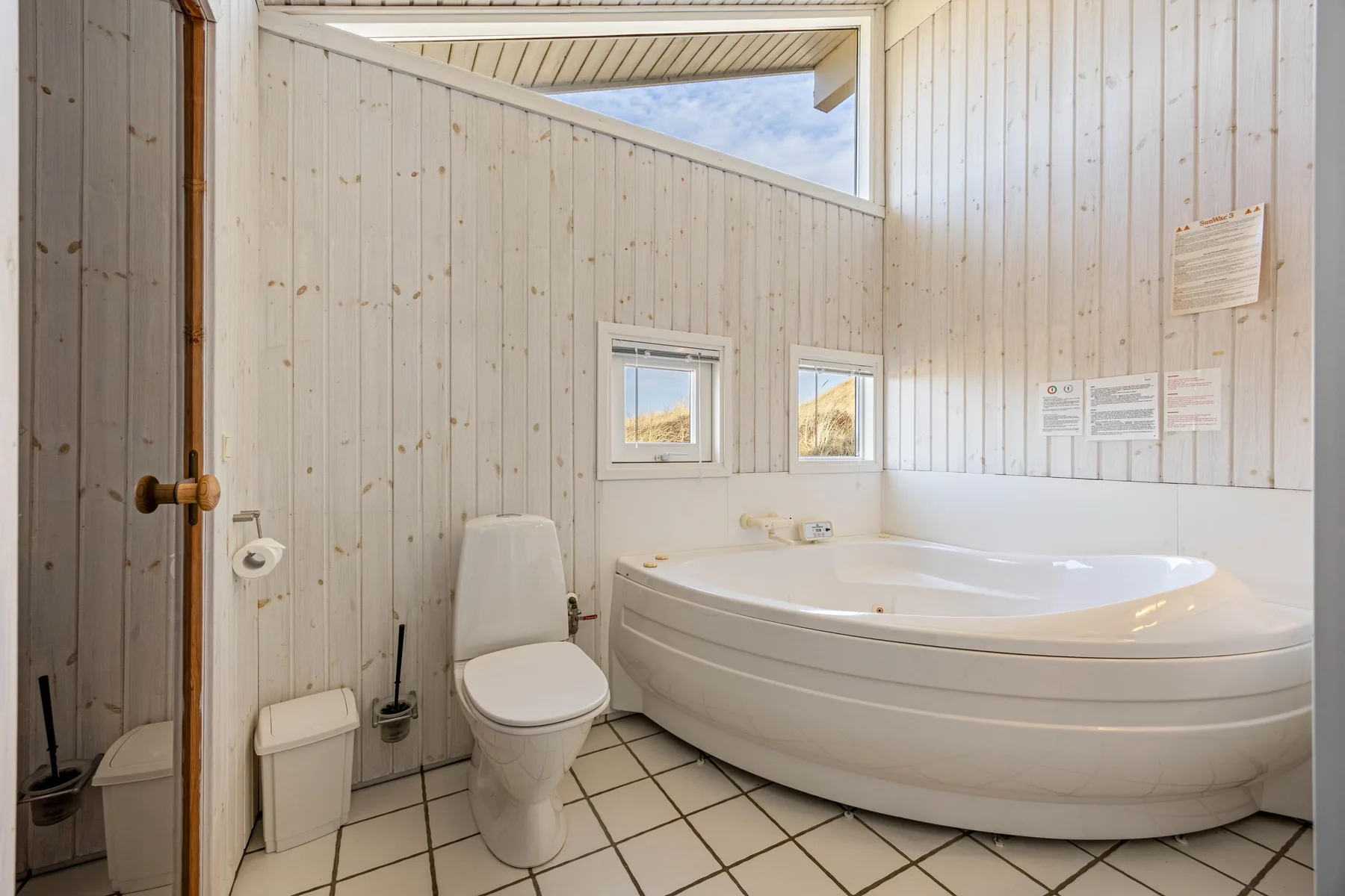 Un baño luminoso con inodoro y una bañera grande bajo ventanas pequeñas.