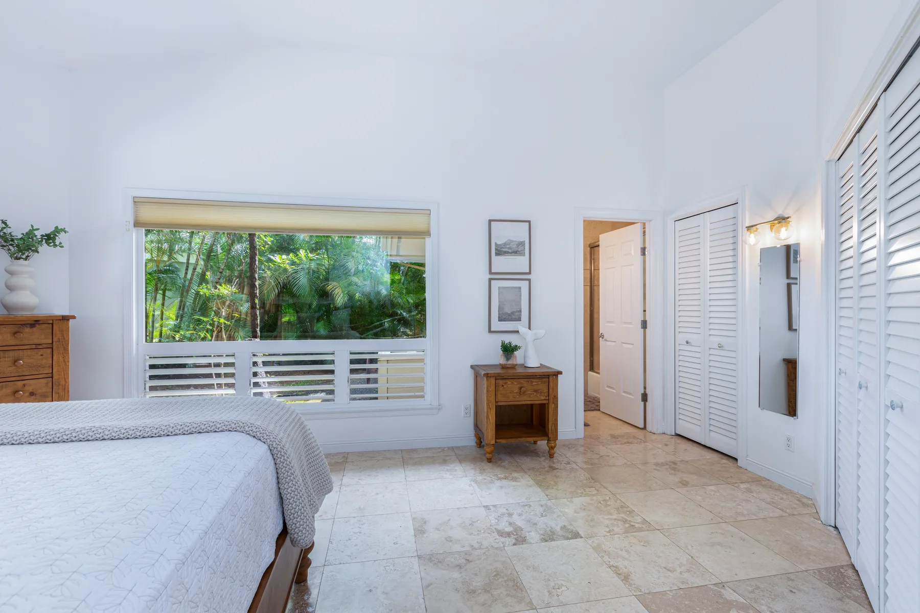 Un dormitorio luminoso con una cama grande, una ventana y vegetación frondosa en el exterior.