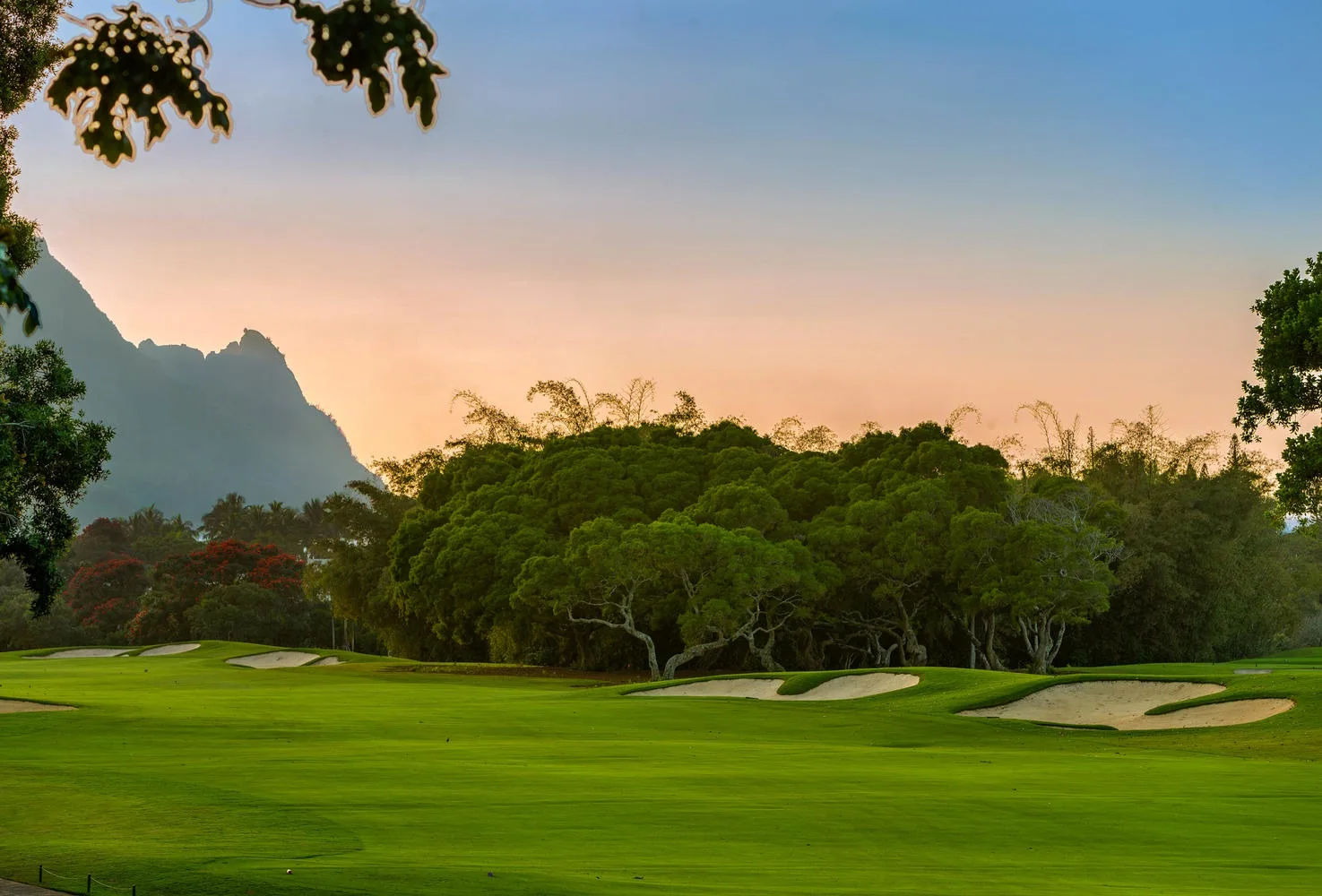 Un campo de golf verde se extiende hacia árboles y montañas bajo un cielo colorido al atardecer.