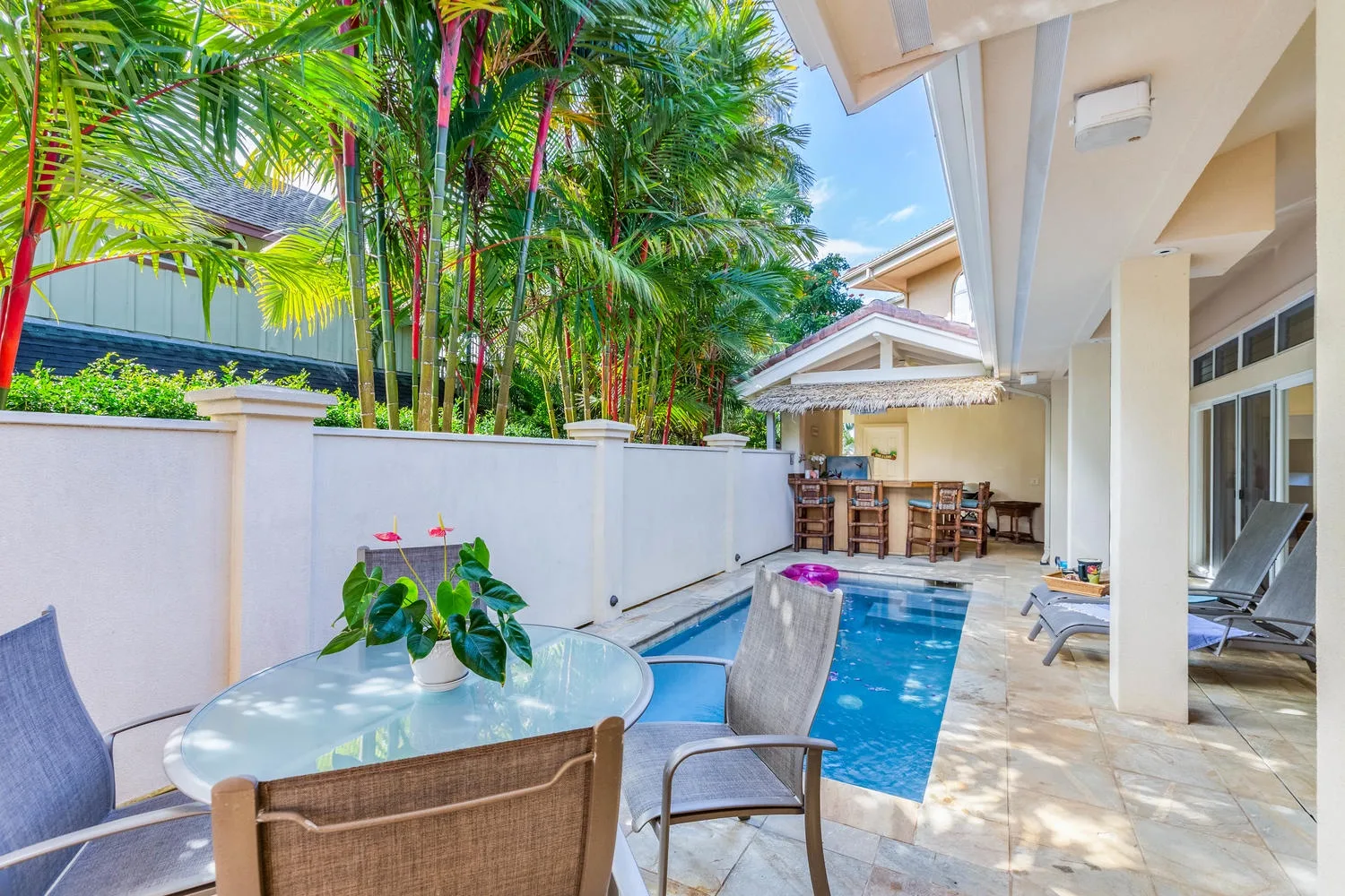 Un patio con piscina, asientos y plantas tropicales junto a una zona de comedor cubierta.