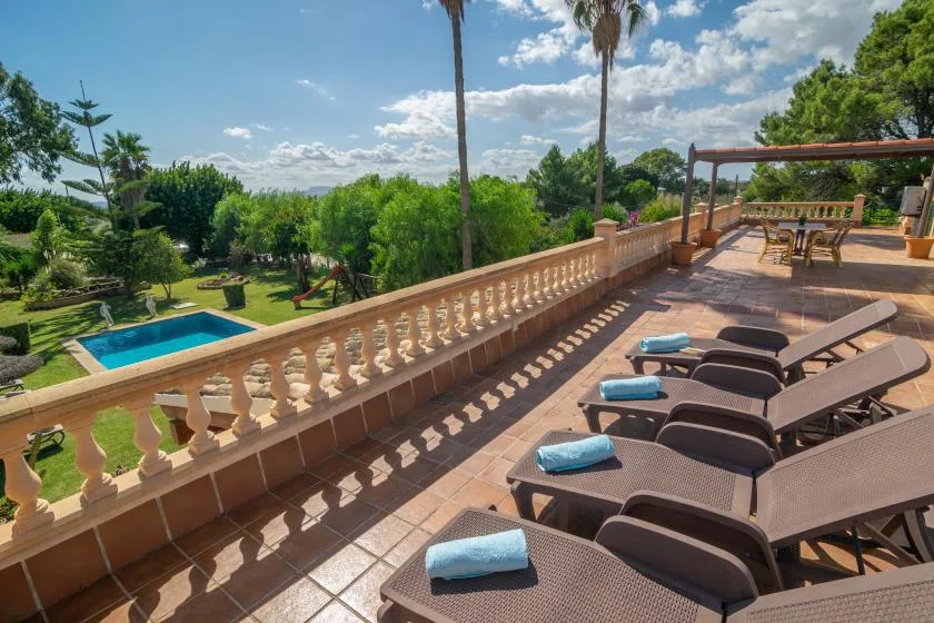 A sunny terrace overlooks a garden and swimming pool, with lounge seating.