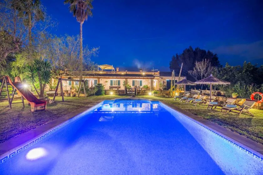 A swimming pool glows blue at night beside a garden and seating area.