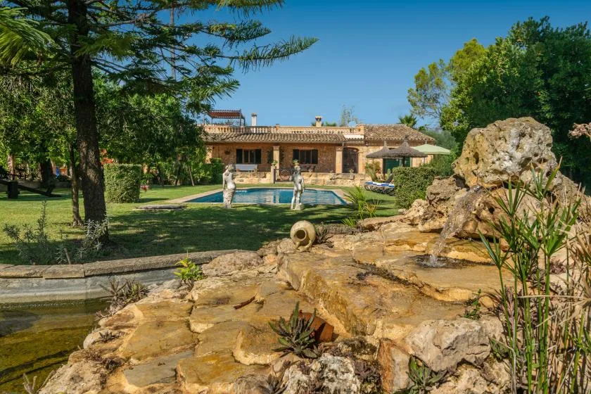 Outdoor garden scene with a rocky water feature, swimming pool, and house in the background.