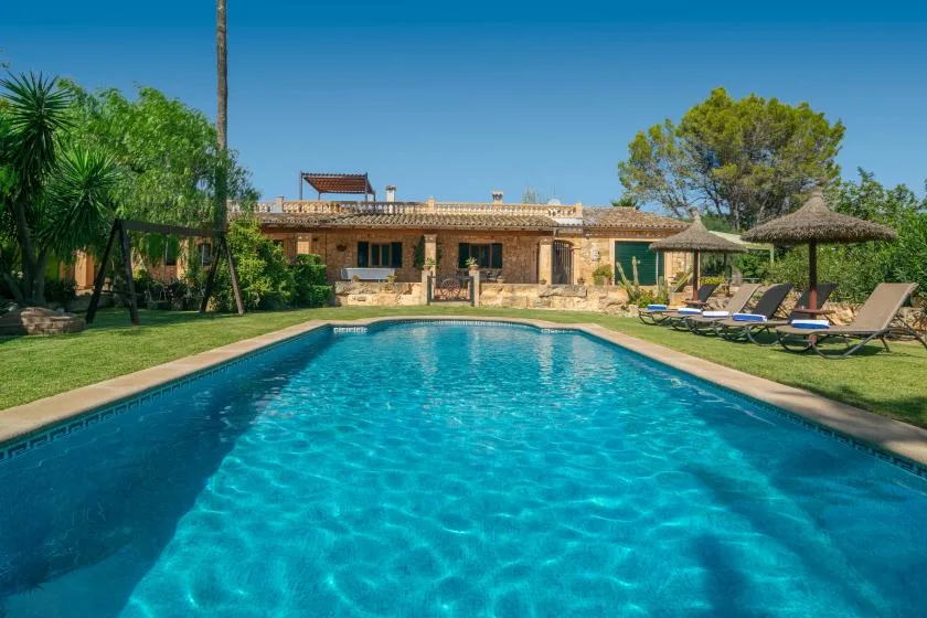 An outdoor swimming pool sits in front of a low stone house and garden.