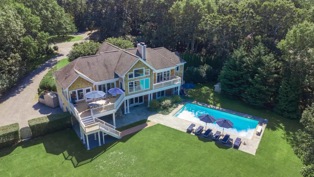 A large house with a swimming pool and outdoor seating beside a wooded area.