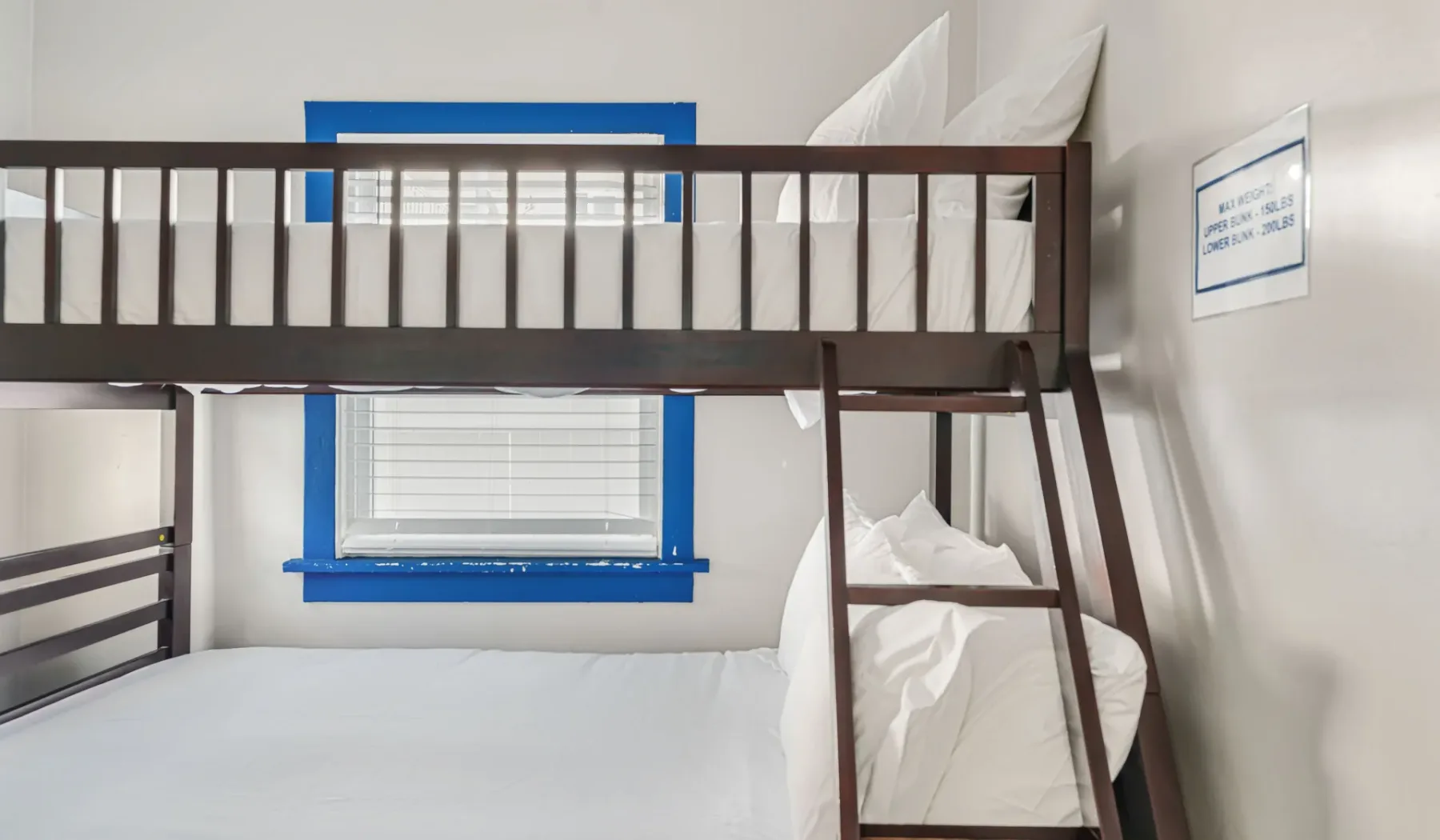 A bunk bed with white bedding stands beside a blue-trimmed window.