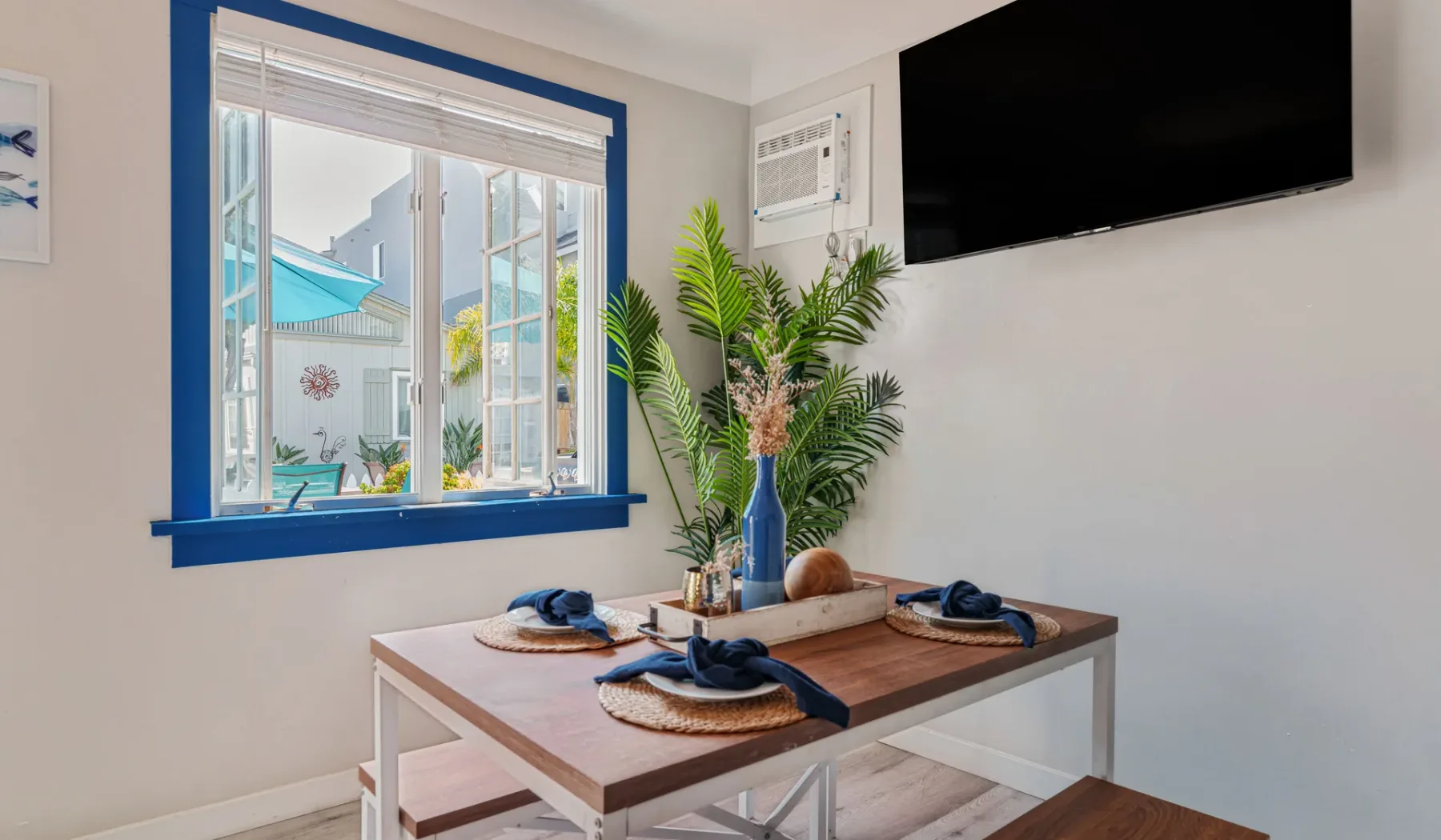 A dining table sits beside a blue-trimmed window, with a television mounted on the wall.