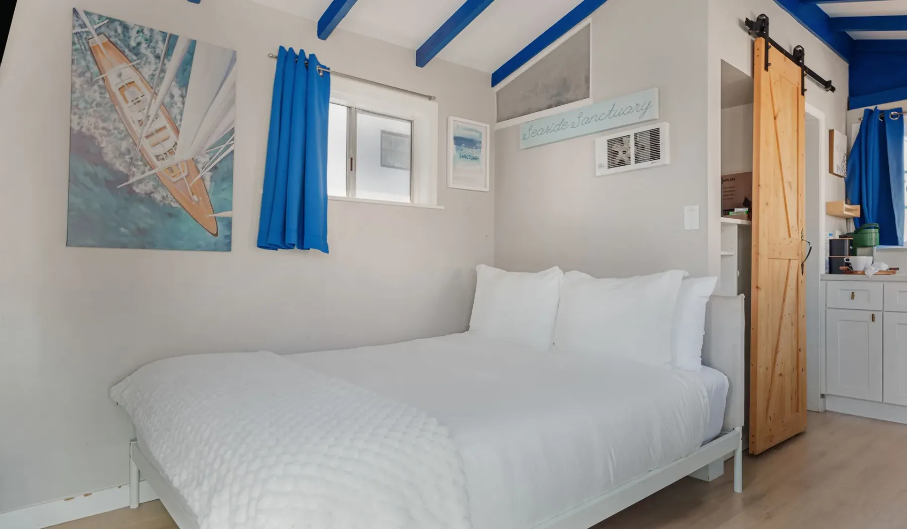 A bedroom with a white bed, blue curtains, and exposed ceiling beams.