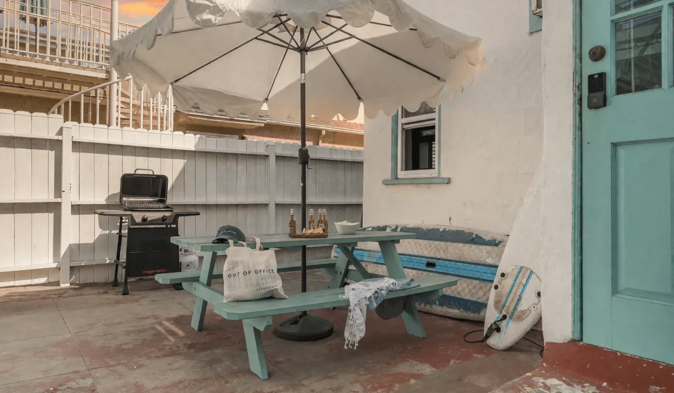 An outdoor patio has picnic-table seating beneath a large umbrella, with a grill nearby.