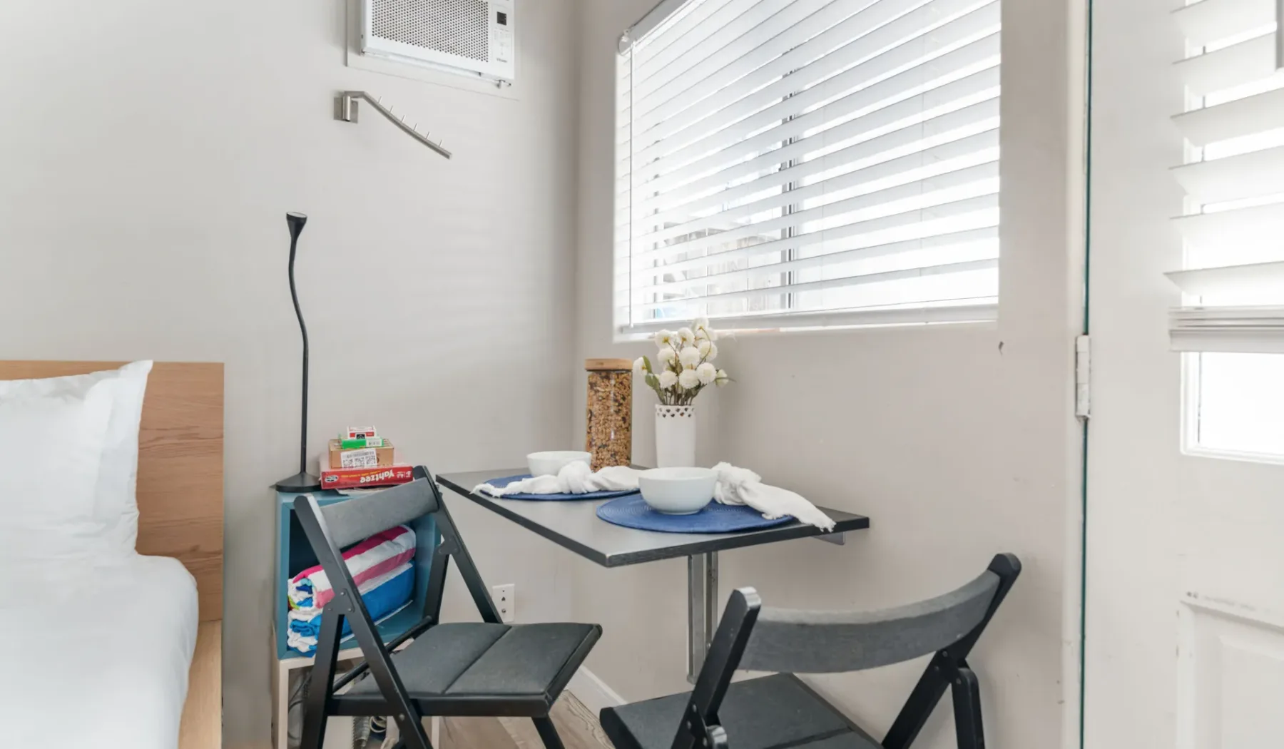 A bright bedroom corner has a small table and folding chairs beside the bed.