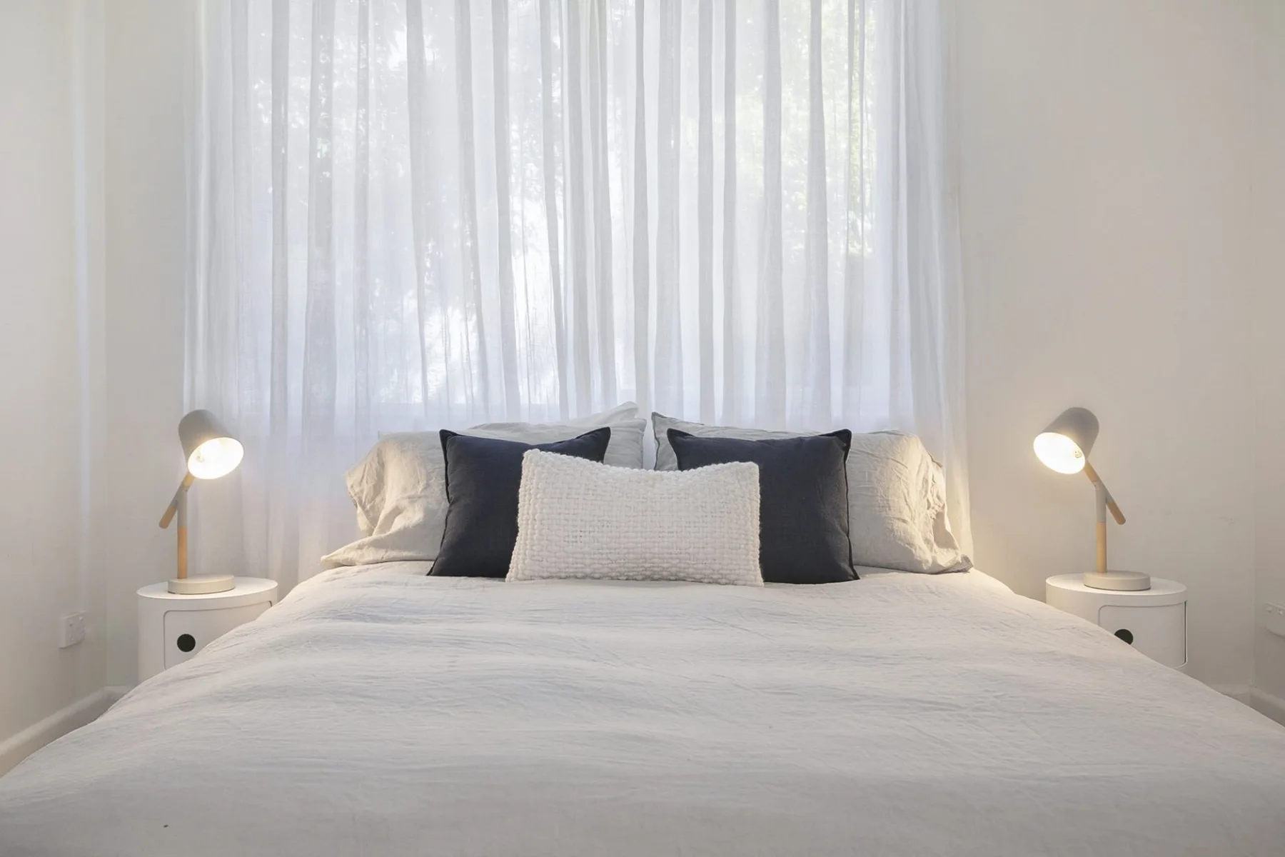 Un dormitorio luminoso con una cama bien arreglada bajo cortinas translúcidas.