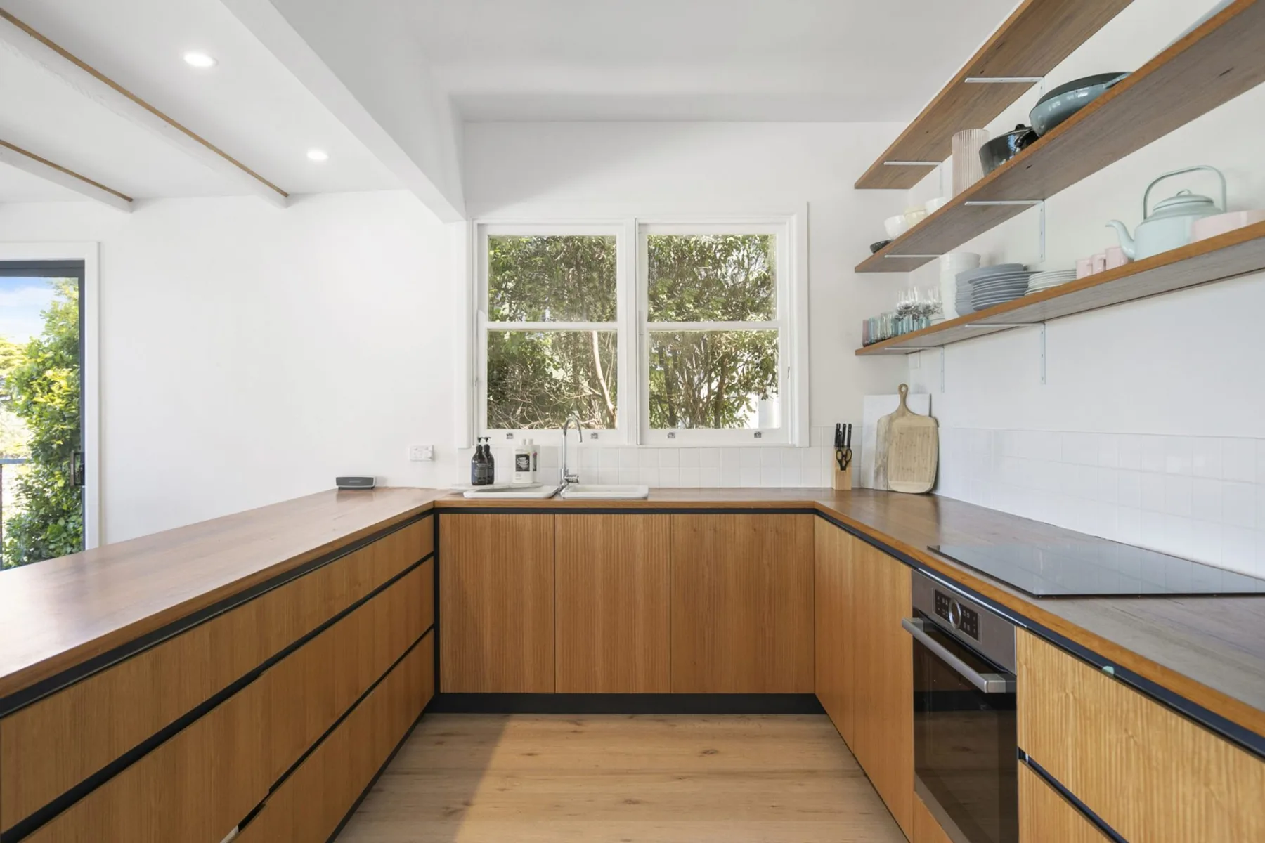 Una cocina moderna con gabinetes de madera, encimeras oscuras y ventanas con vista a los árboles.