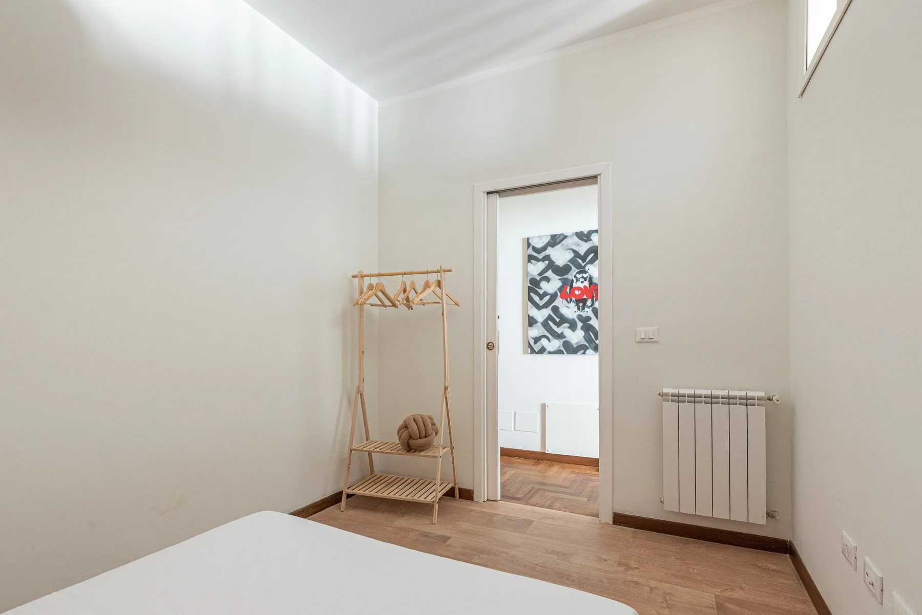 A bright bedroom with a wooden clothes rack and a doorway to another room.