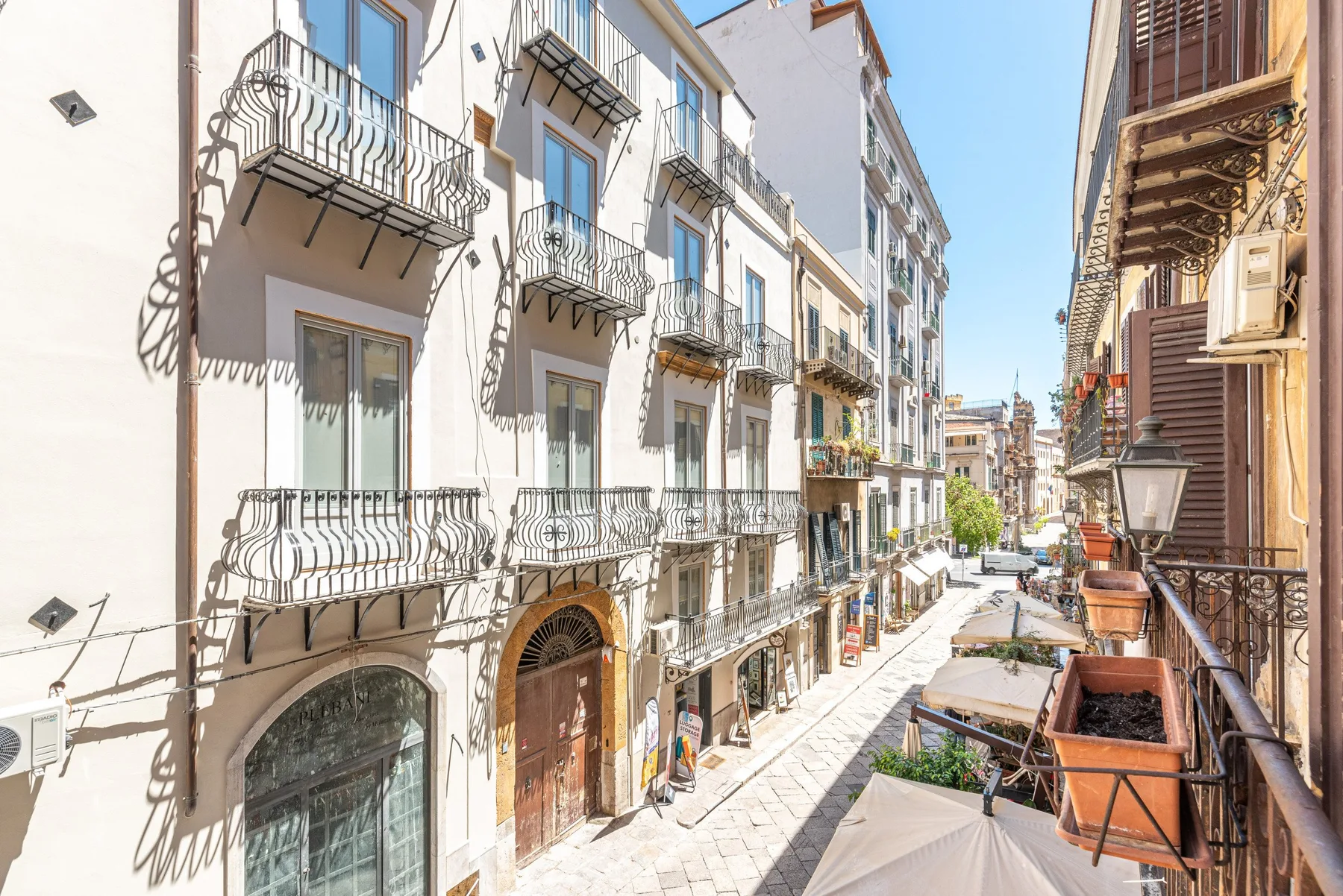 A sunlit pedestrian street runs between buildings with balconies and outdoor seating.