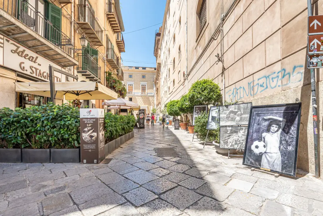 A narrow pedestrian street lined with café seating, potted plants, and display boards.