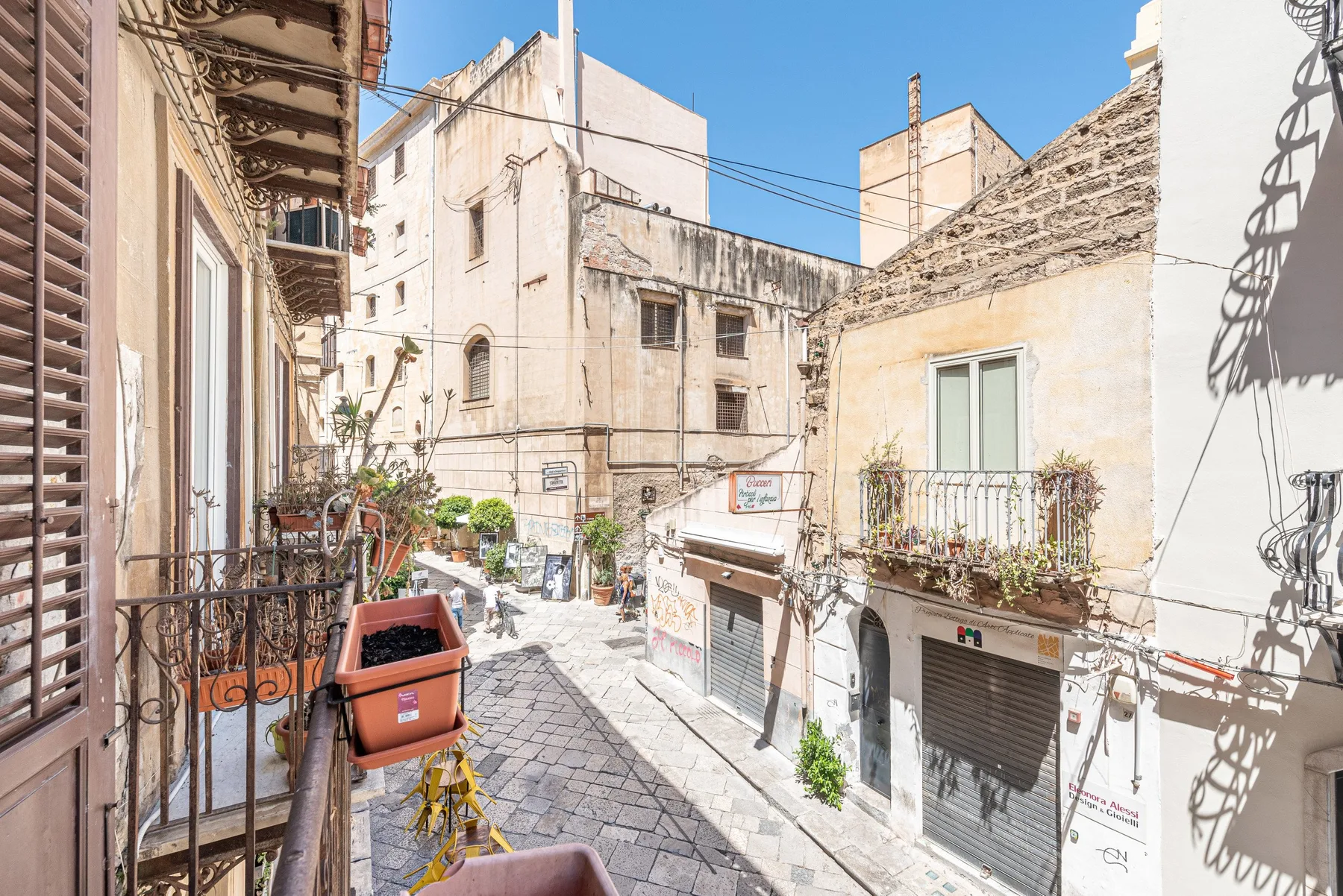 A narrow paved street runs between closely spaced buildings with balconies and potted plants.