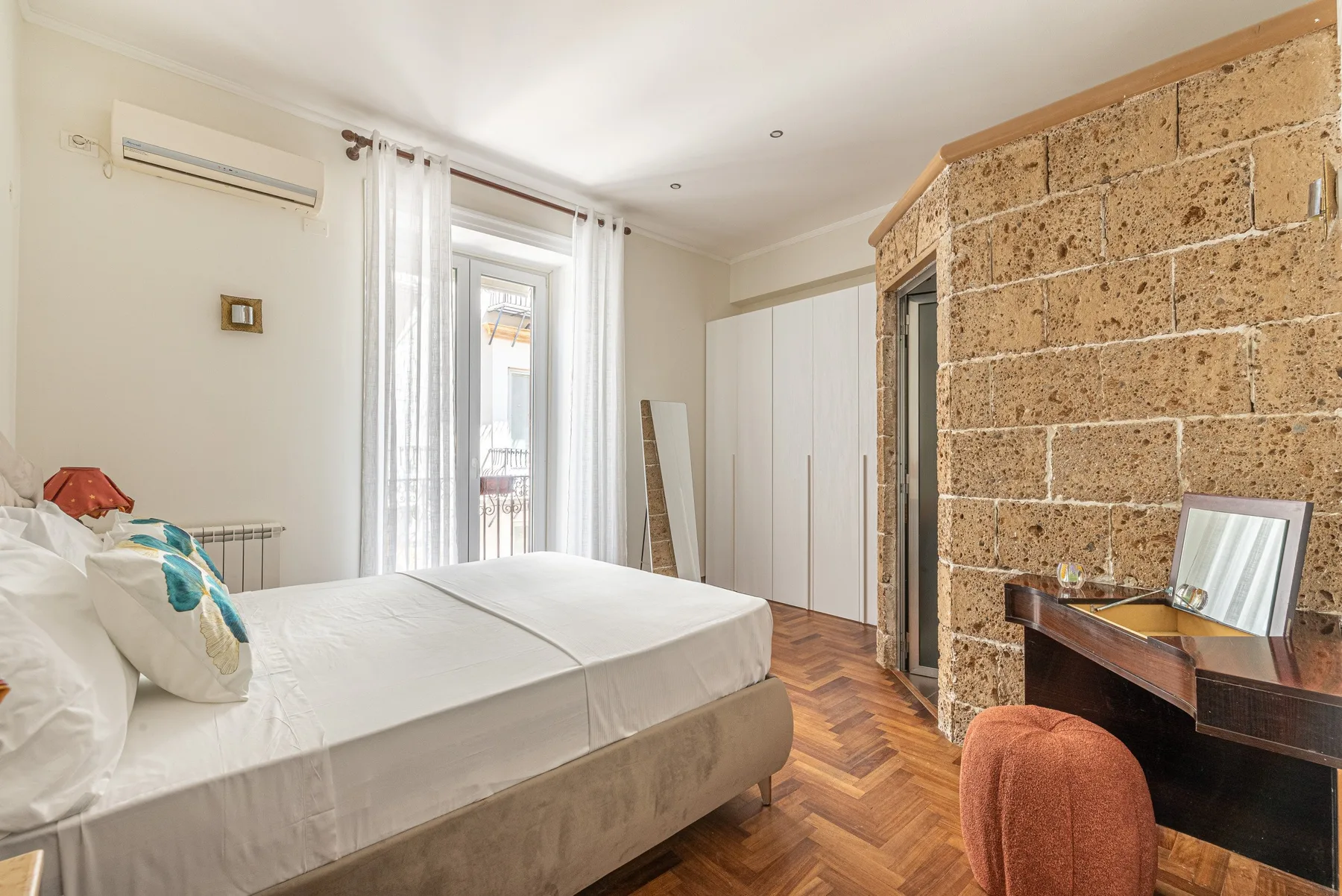 A bedroom with a bed, tall windows, and a stone-textured wall.