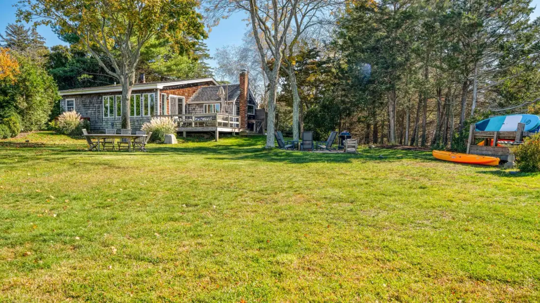 A house with a wooden deck sits beside a broad grassy yard and trees.