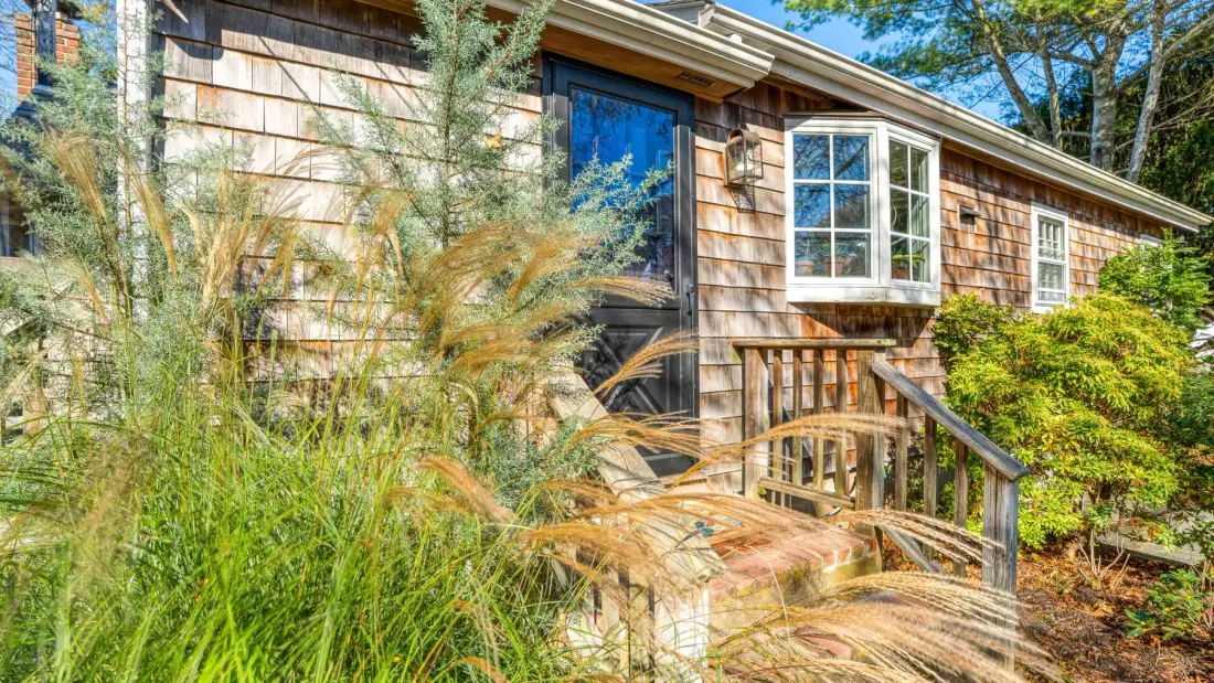 A wood-sided house exterior with a small entry porch, surrounded by greenery.