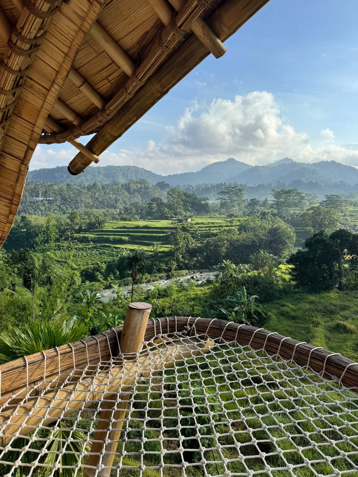 A rope net hammock overlooks green fields and distant hills beneath a thatched roof.