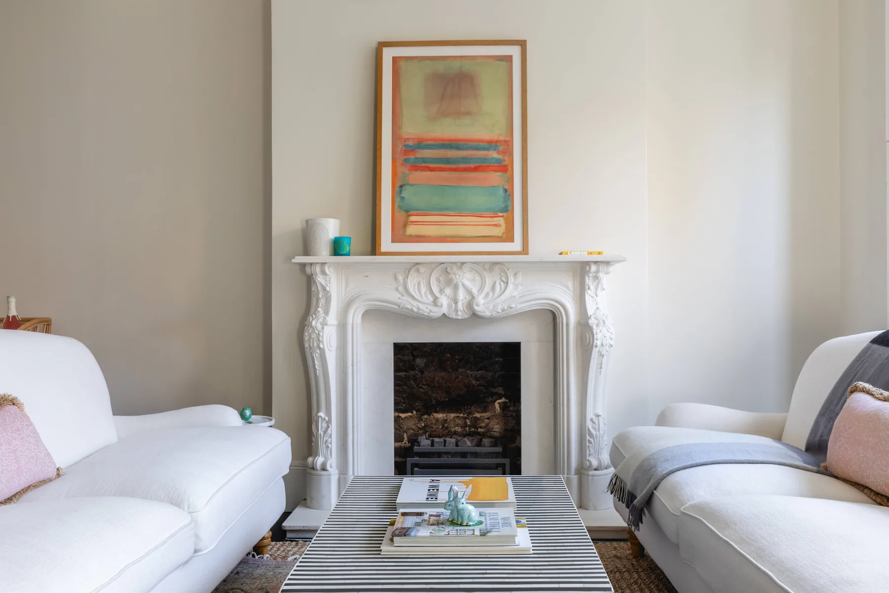 Una sala luminosa con asientos frente a una chimenea ornamentada y una obra enmarcada.