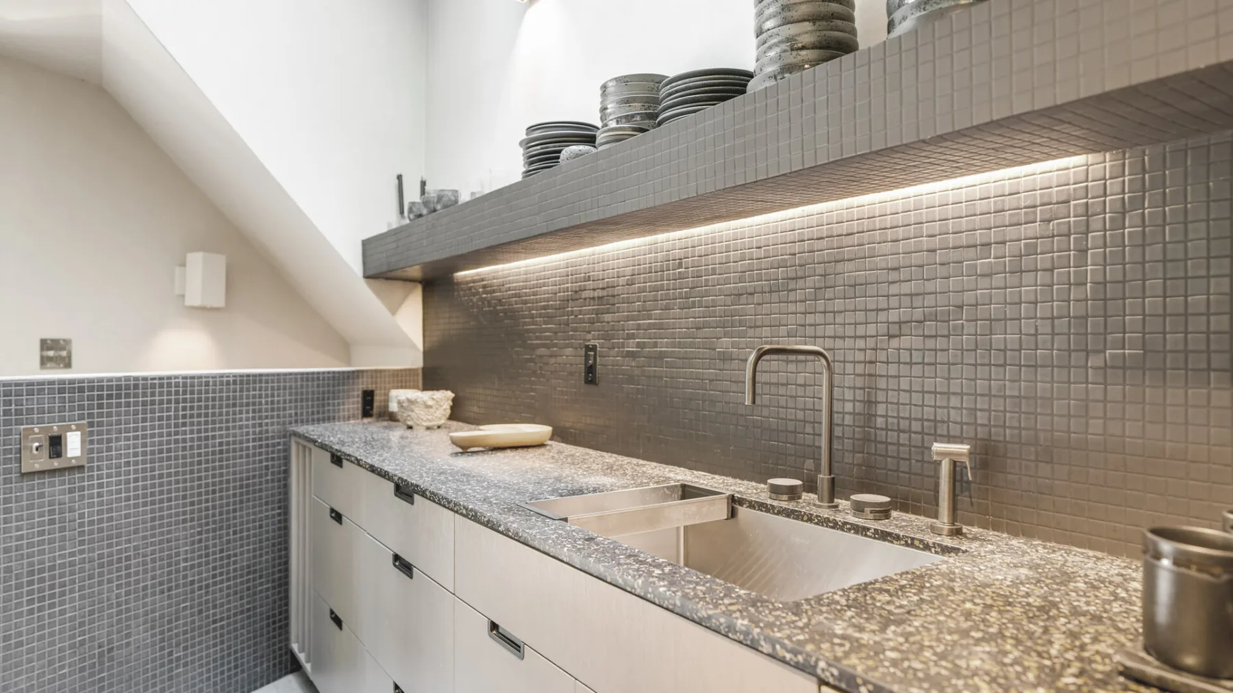 Una encimera de cocina revestida de azulejos, con fregadero doble y platos en el estante superior.
