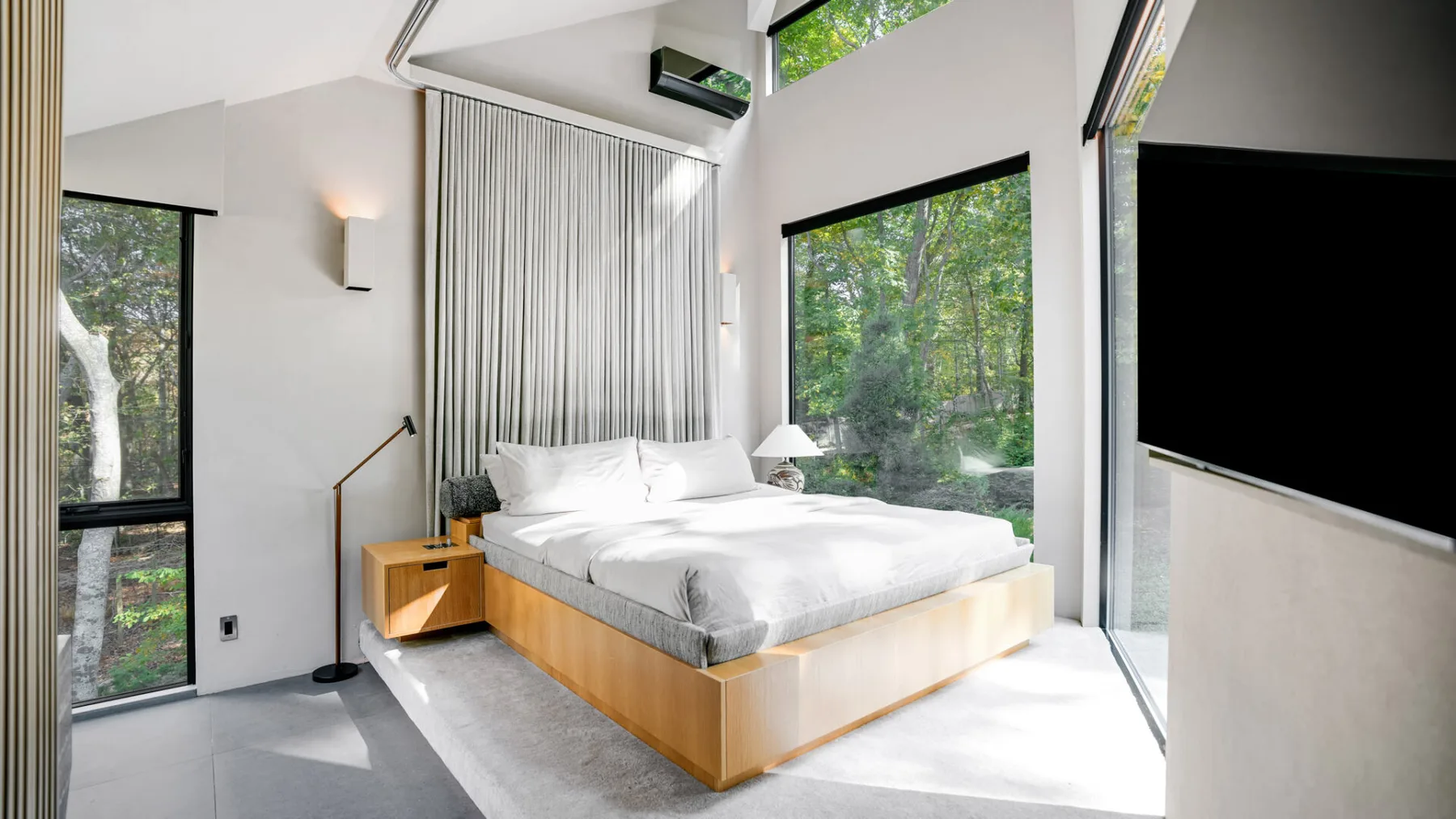 Un dormitorio luminoso con una cama baja junto a grandes ventanas con vistas a los árboles.