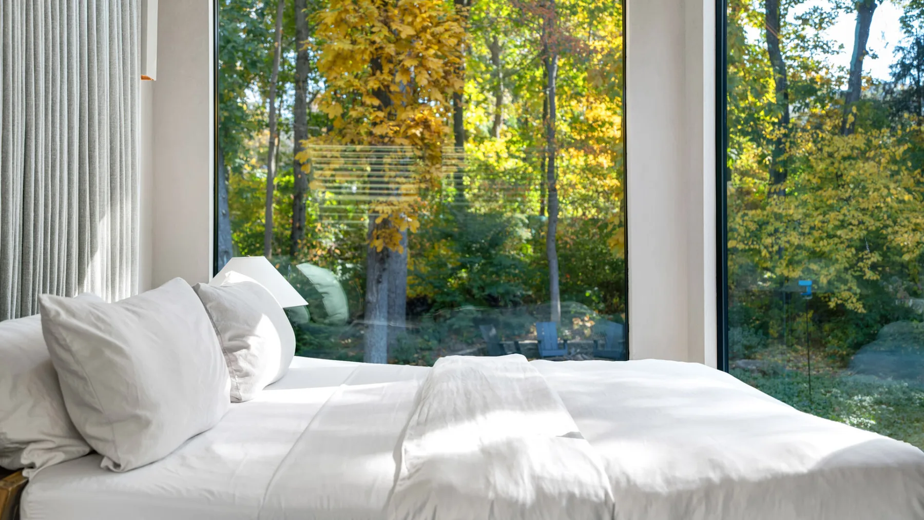 Un dormitorio luminoso con ropa de cama blanca junto a grandes ventanas con vistas a árboles frondosos.