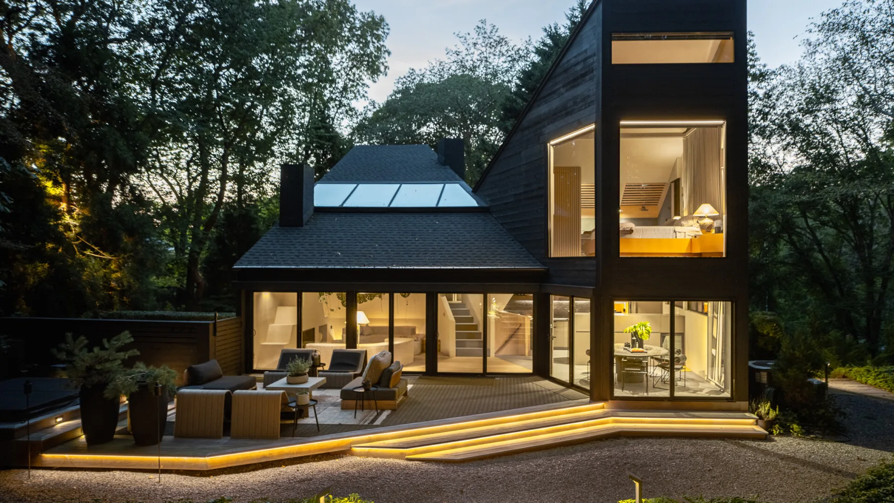 Una casa moderna se ilumina al anochecer, con grandes ventanales junto a una terraza.