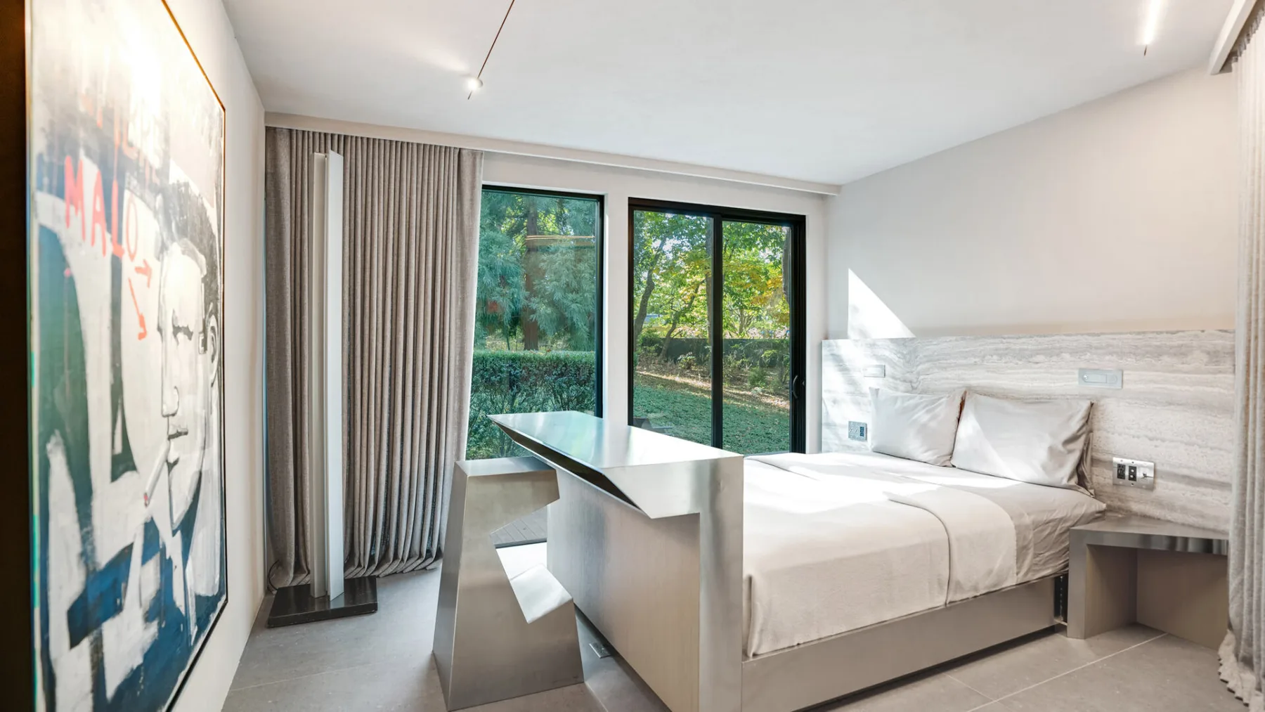Un dormitorio con una cama, un escritorio y grandes ventanas con vistas a la vegetación.
