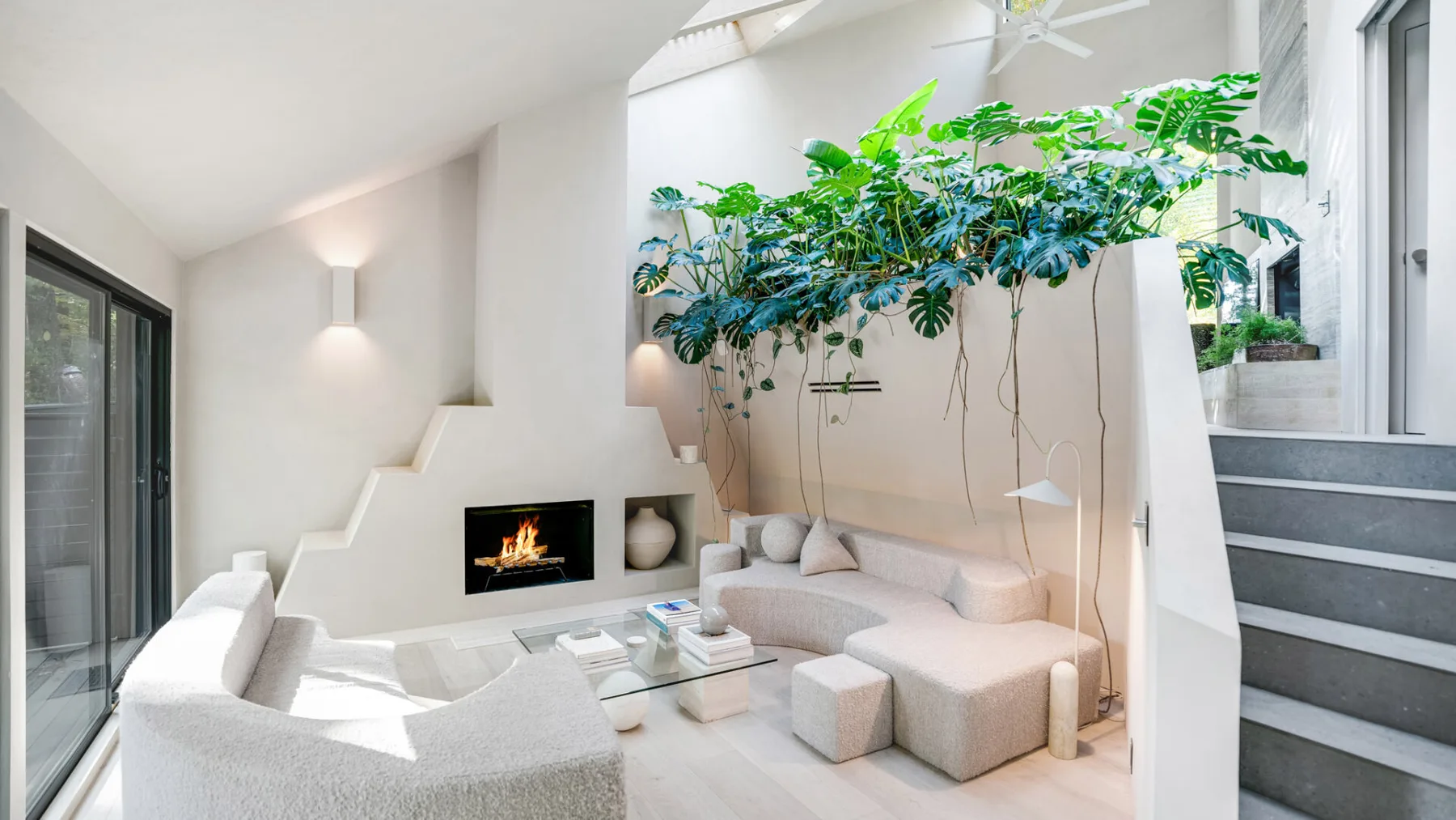 Una sala luminosa con asientos bajos, una chimenea y plantas frondosas.