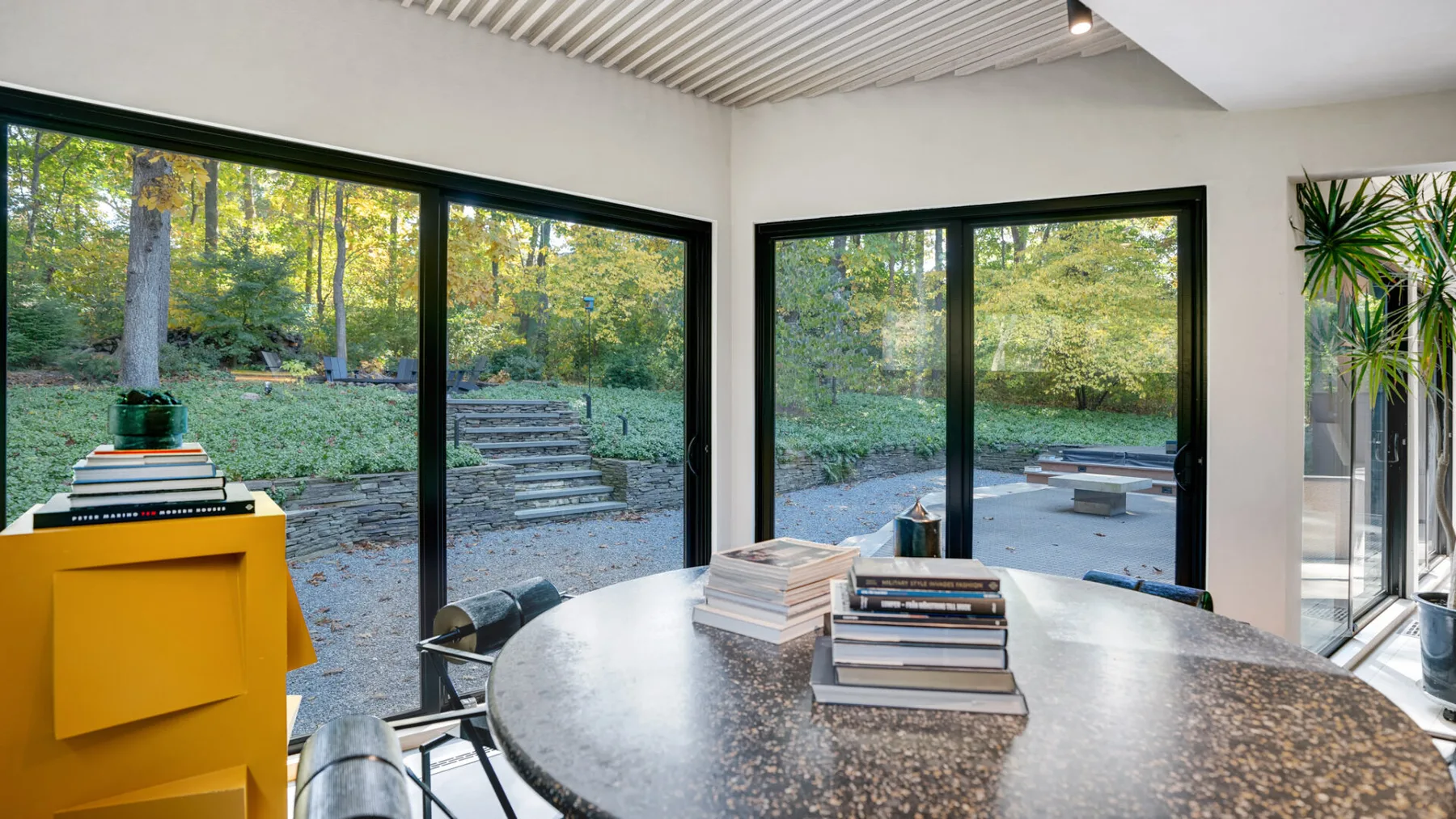 Una habitación luminosa con una mesa redonda junto a grandes ventanales que dan a un jardín arbolado.