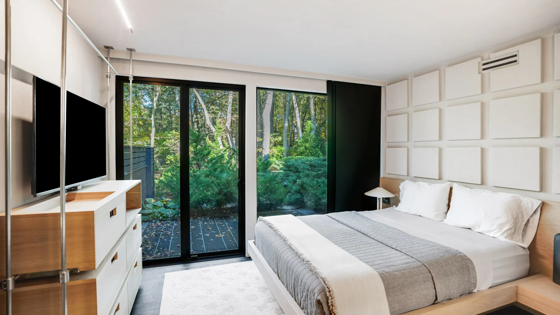 Un dormitorio luminoso con una cama tendida y grandes ventanas con vistas a la vegetación.