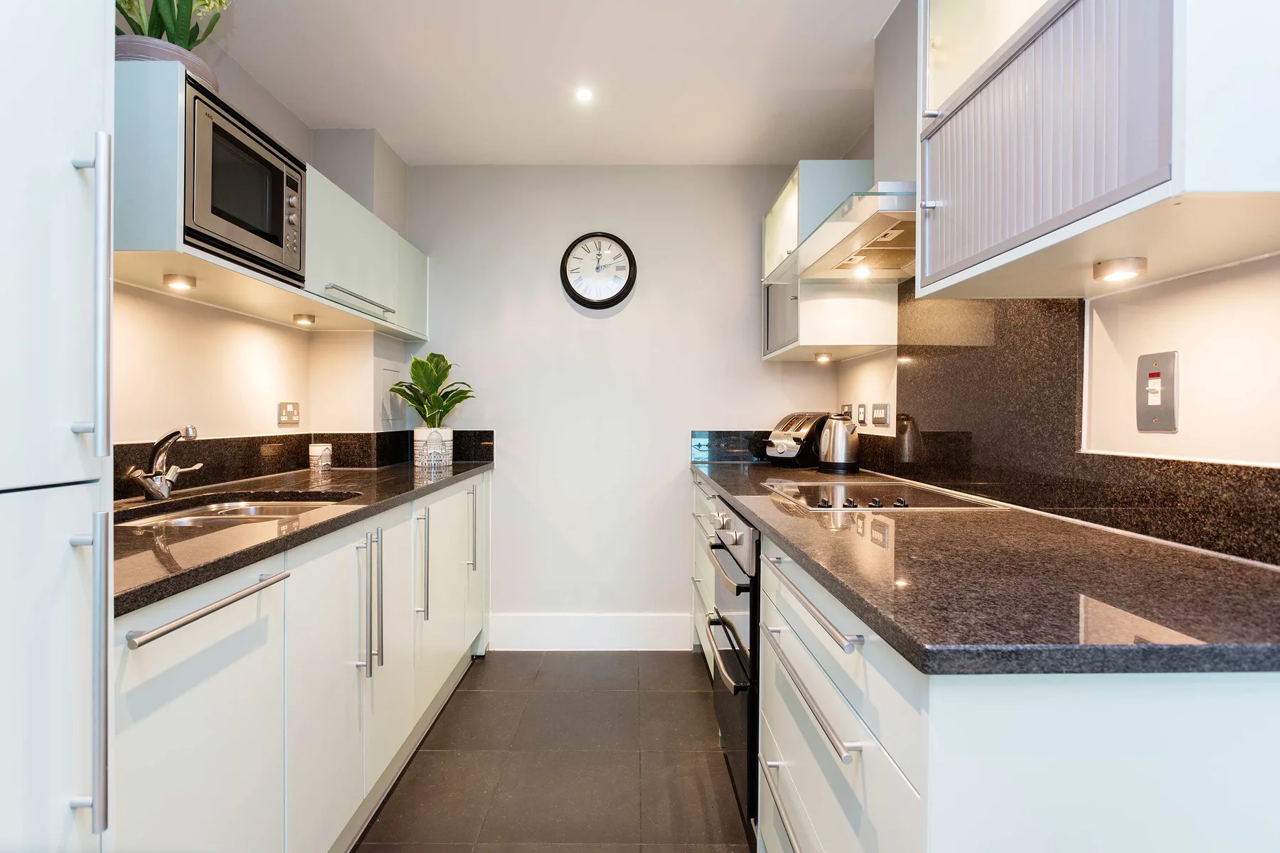 A modern kitchen with parallel counters, pale cabinets, and a clock on the back wall.