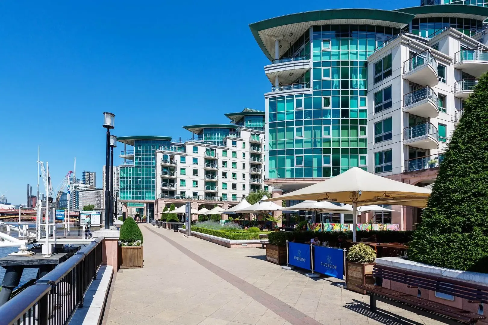 A riverside promenade passes modern glass-fronted buildings and outdoor seating.