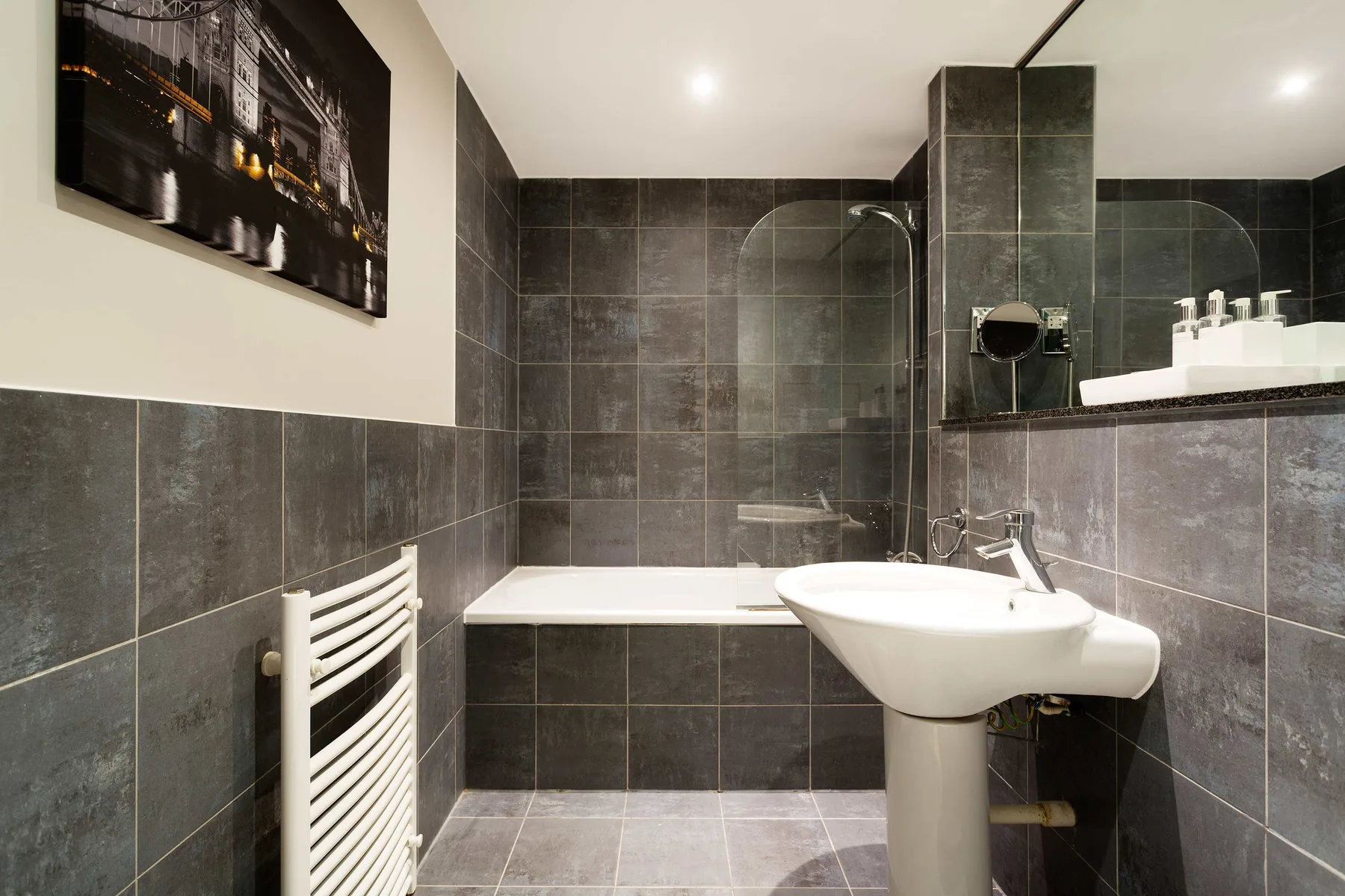 A tiled bathroom with a bathtub, pedestal sink, and wall-mounted towel warmer.