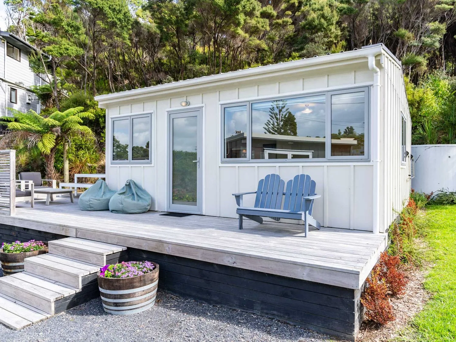 Una pequeña cabaña blanca con terraza de madera y asientos al aire libre entre árboles.
