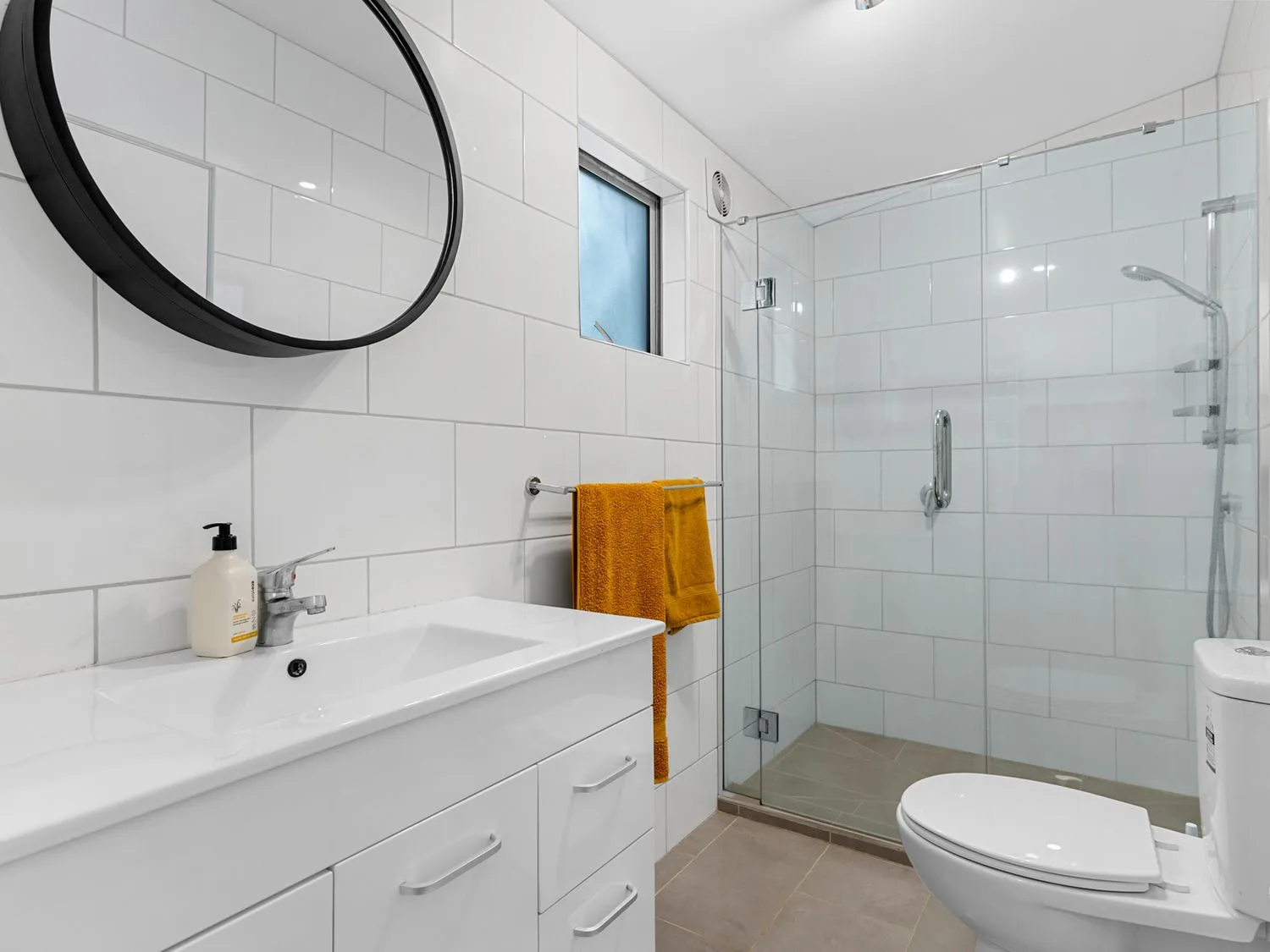 Un baño con azulejos, lavabo, espejo redondo, inodoro, ducha de vidrio y toalla naranja.