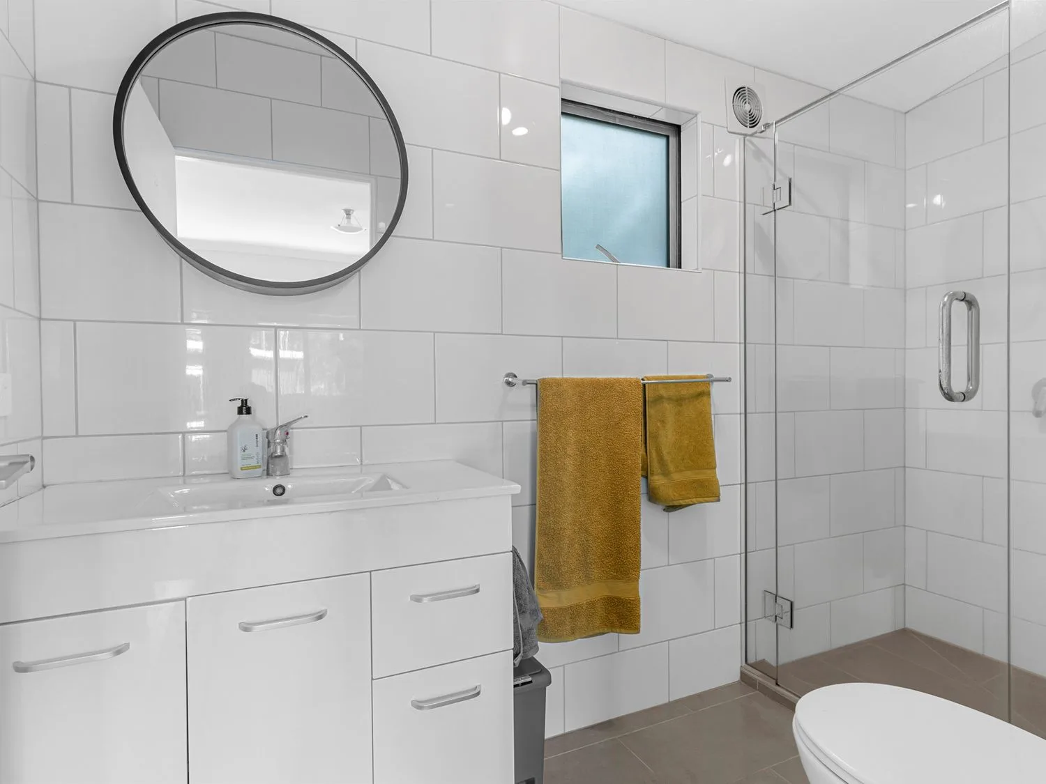 Un baño con un espejo redondo sobre un mueble de lavabo blanco, toallas amarillas en un toallero y una ducha con puerta de vidrio en Pearls Embrace Mangawhai Heads Studio, en Mangawhai Heads