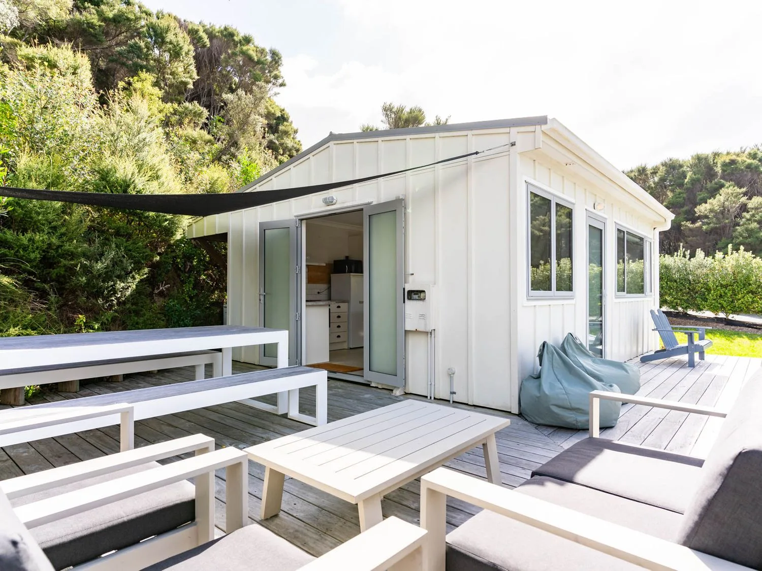 Una terraza exterior de madera con muebles blancos para sentarse, una mesa de pícnic y pufs junto a un pequeño edificio blanco en Pearls Embrace Mangawhai Heads Studio, en Mangawhai Heads