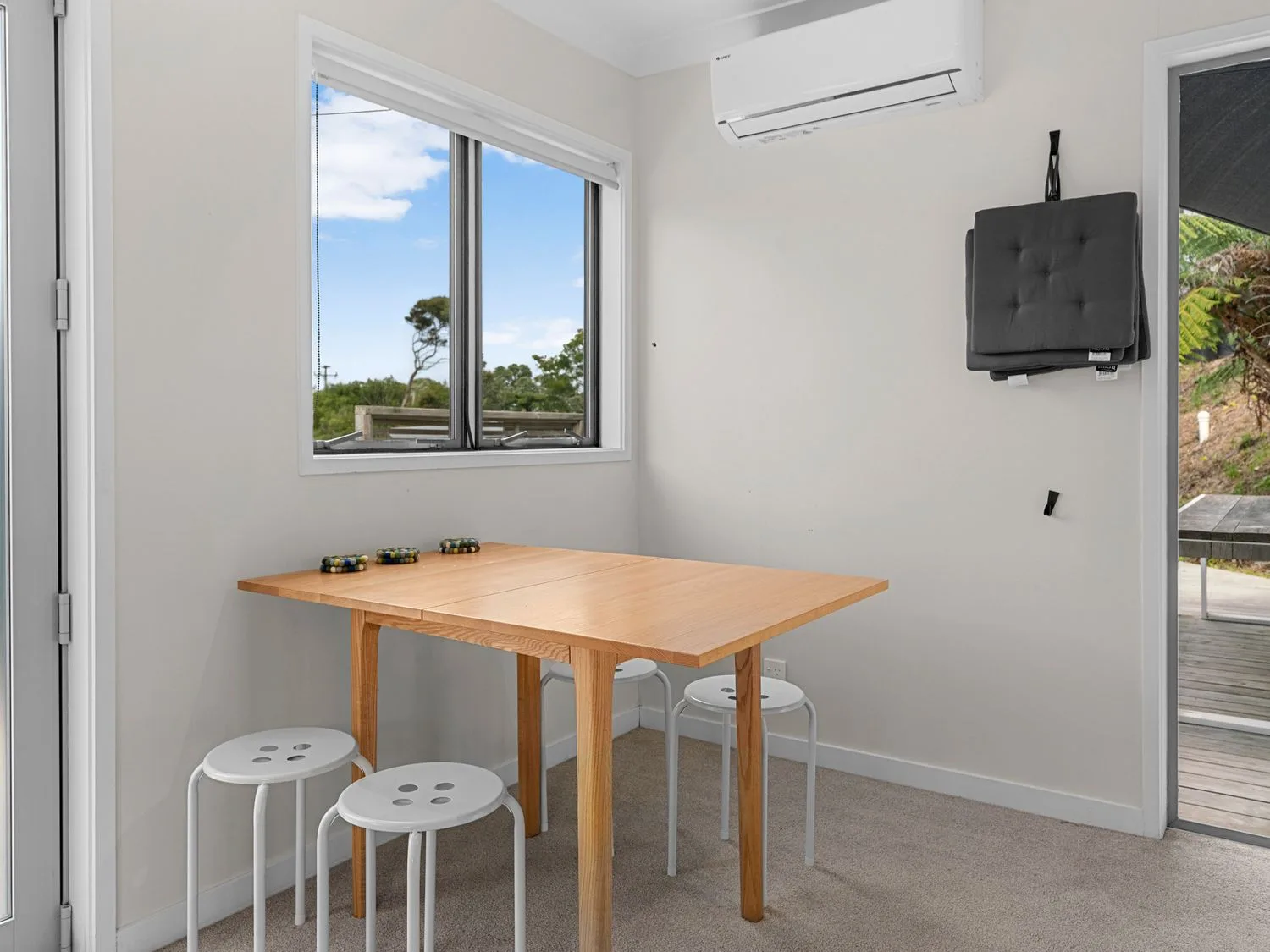 Una zona de comedor con una mesa de madera, cuatro taburetes blancos y una ventana con vistas a la vegetación exterior en Pearl's Embrace - Mangawhai Heads Studio, en Mangawhai Heads